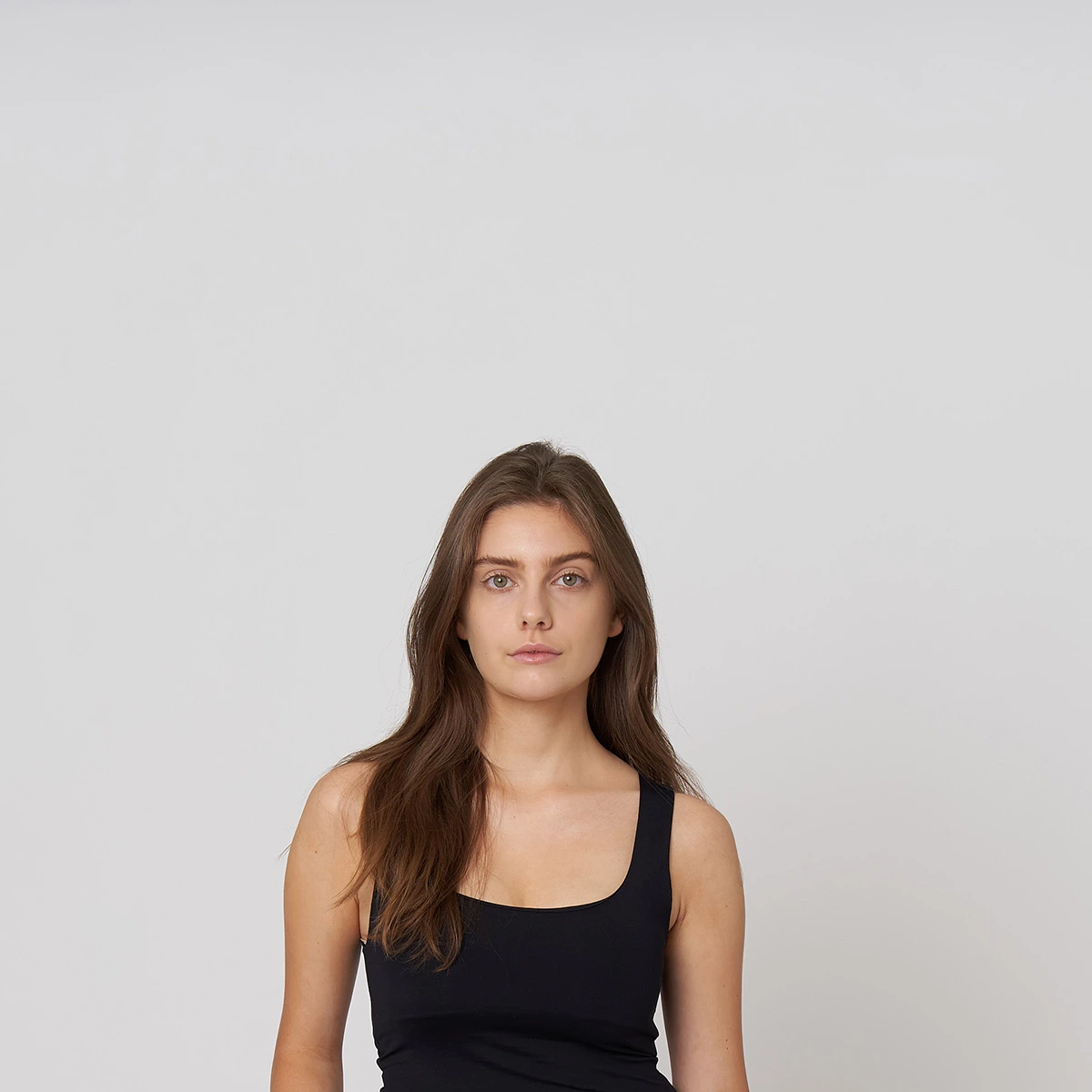 Une femme aux longs cheveux bruns, vêtue d'un haut noir sans manches, pose sur une toile de fond gris clair, face à l'appareil photo, pour une prise de tête en studio.