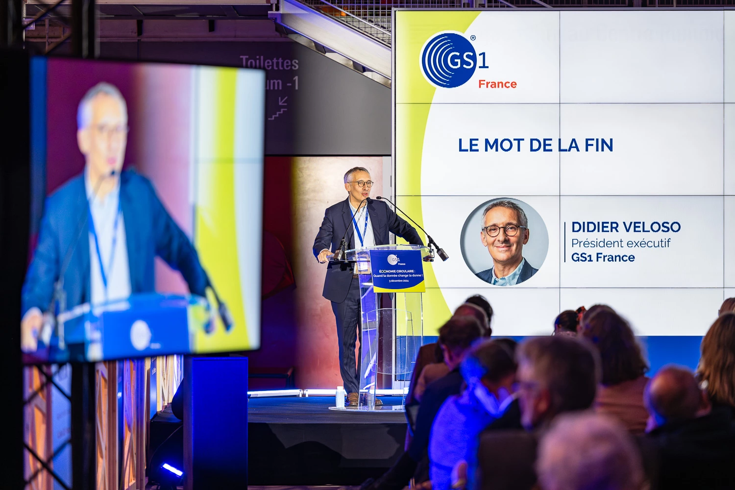 Un orateur sur un podium s'adresse à un public, avec en toile de fond une projection montrant le nom et la photo de GS1 France et de Didier Veloso.