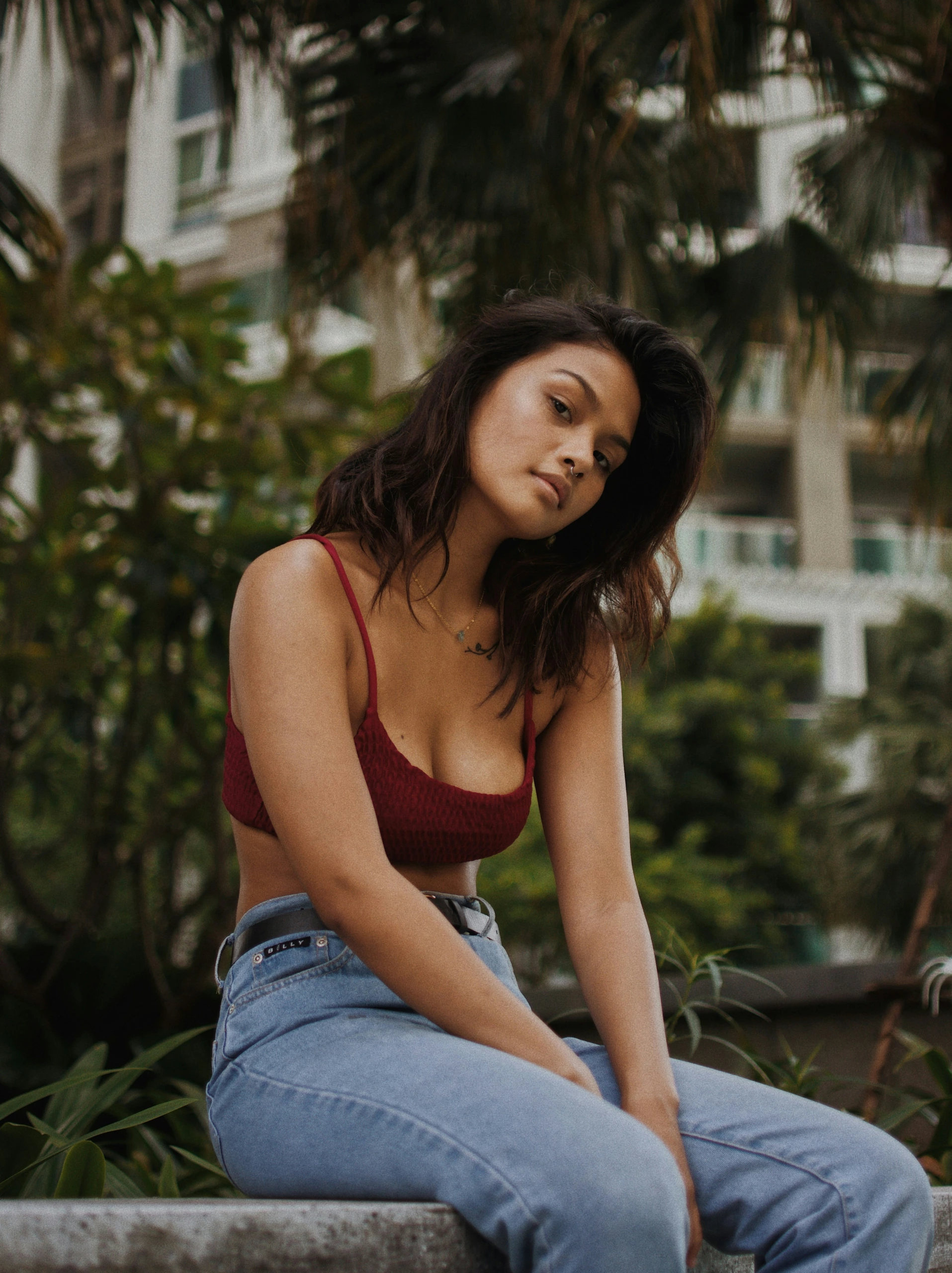 Un mannequin aux cheveux bruns mi-longs, vêtu d'un crop top rouge et d'un jean bleu, pose en plein air sur du béton, près d'une verdure et d'un bâtiment, pour une séance de photos de mode.
