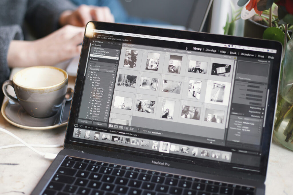 Un MacBook Pro équipé d'un logiciel d'édition Adobe affiche des vignettes de photos en noir et blanc ; en arrière-plan, les mains d'une personne actionnent un équipement.
