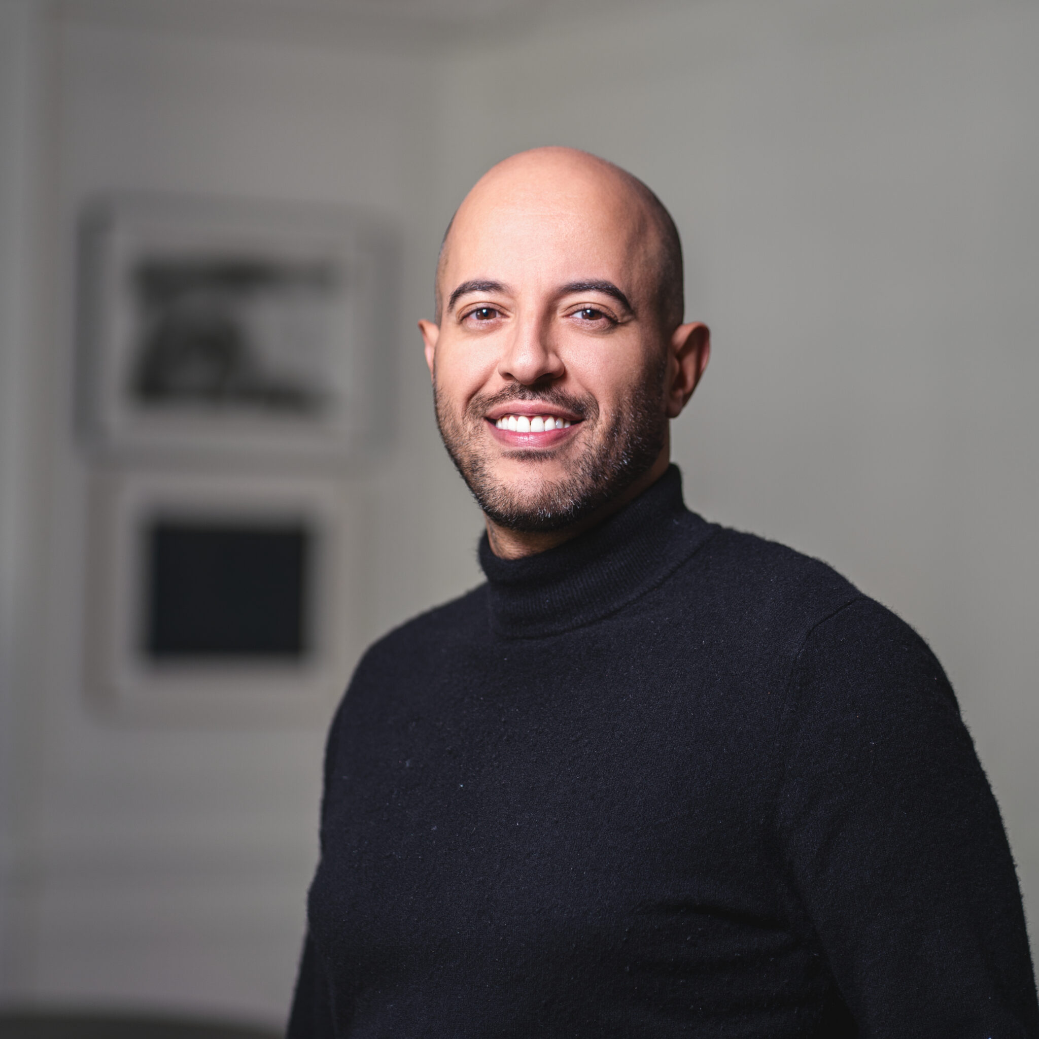 Portrait professionnel d'un homme au crâne rasé et à la barbe taillée, vêtu d'un col roulé noir, dans un bureau parisien, sur fond d'art mural flou.