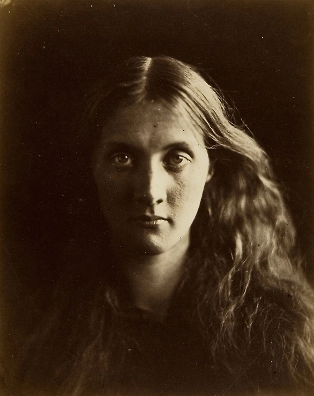 Portrait de studio en tons sépia d'une femme aux cheveux longs, ondulés et coiffés au centre, faisant face à l'appareil photo sur un fond uni et sombre.