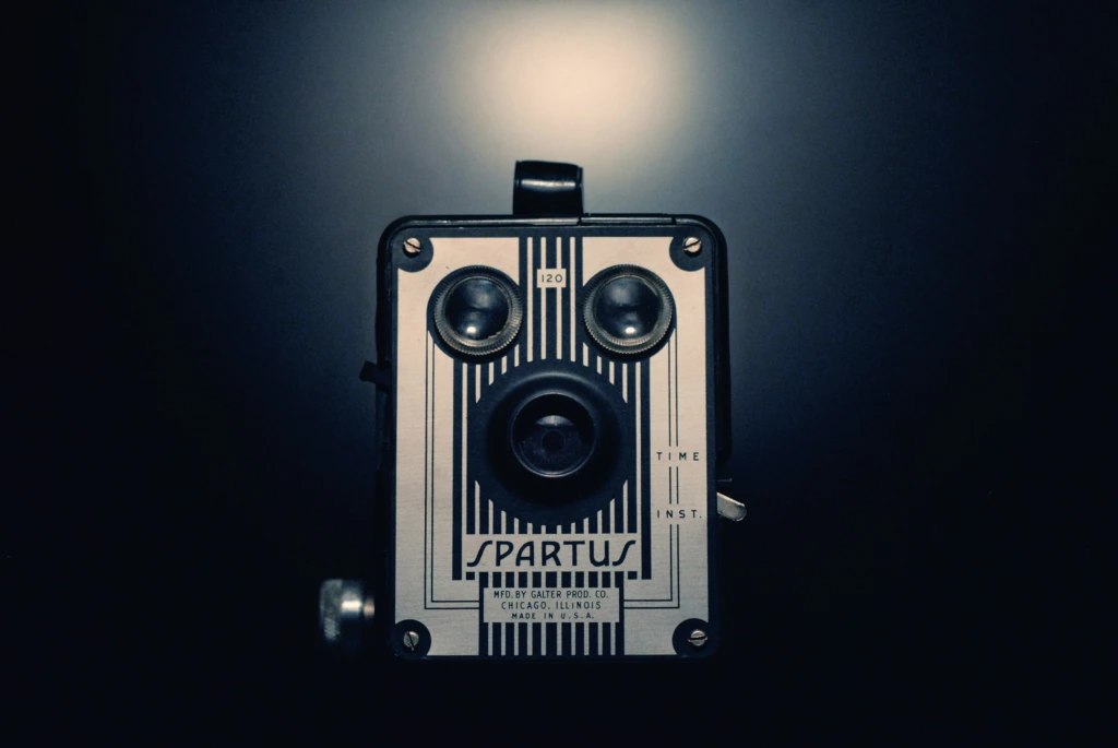 Un appareil photo Spartus vintage, éclairé par un projecteur sur un fond sombre, présente sa façade métallique rayée, ses deux objectifs et ses commandes étiquetées.