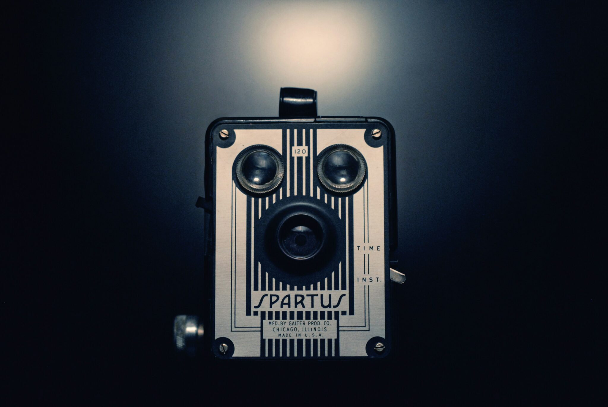 Un appareil photo Spartus vintage, éclairé par un projecteur sur un fond sombre, présente sa façade métallique rayée, ses deux objectifs et ses commandes étiquetées.