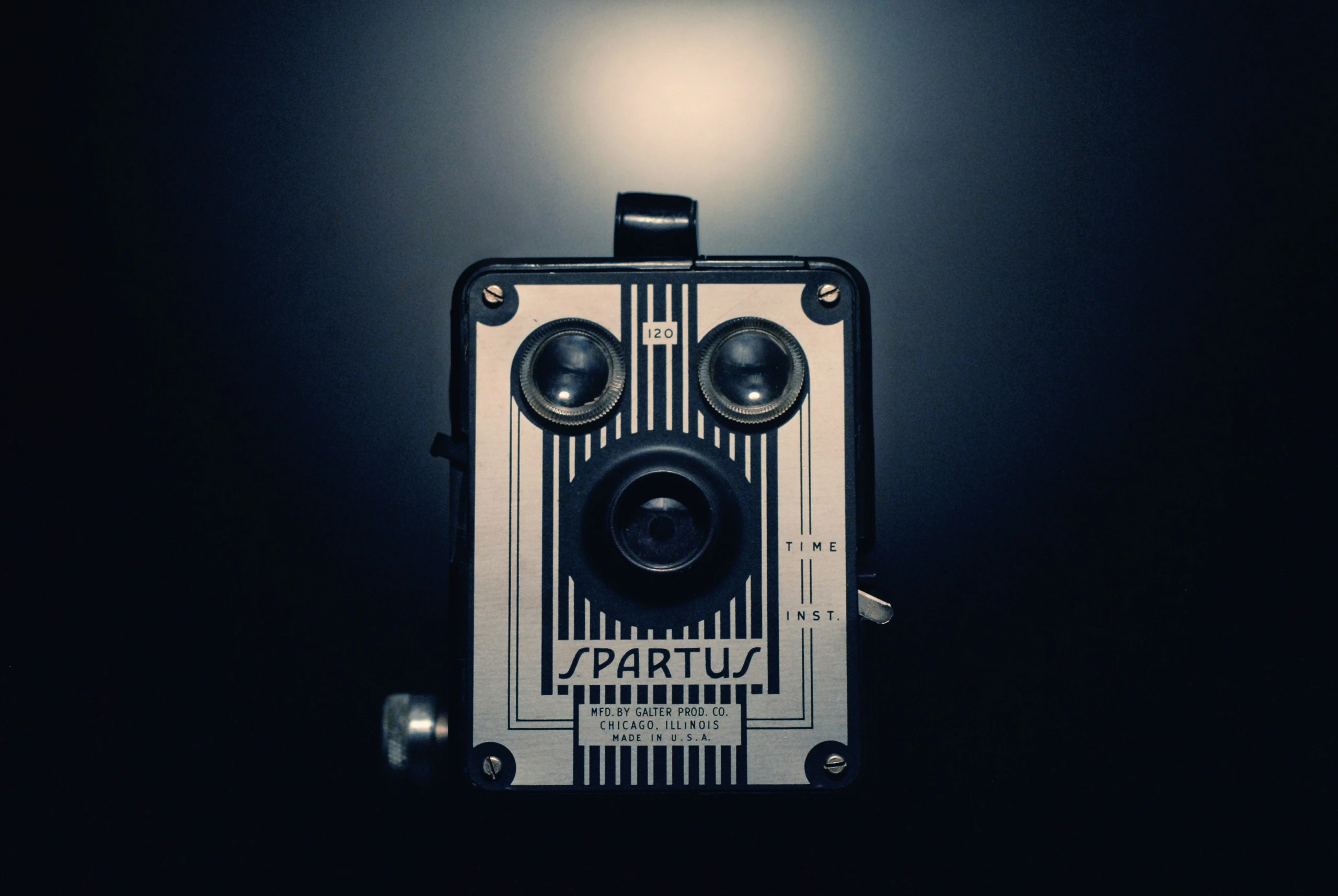 Un appareil photo Spartus vintage, éclairé par un projecteur sur un fond sombre, présente sa façade métallique rayée, ses deux objectifs et ses commandes étiquetées.
