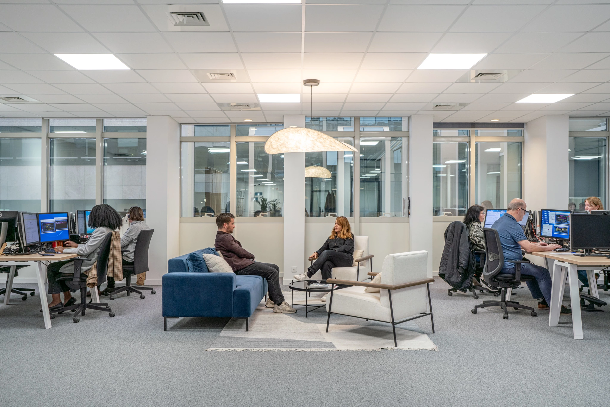 Espace de travail professionnel capturé en haute résolution : personnel devant des ordinateurs, espace central pour s'asseoir, éclairage naturel et artificiel abondant.