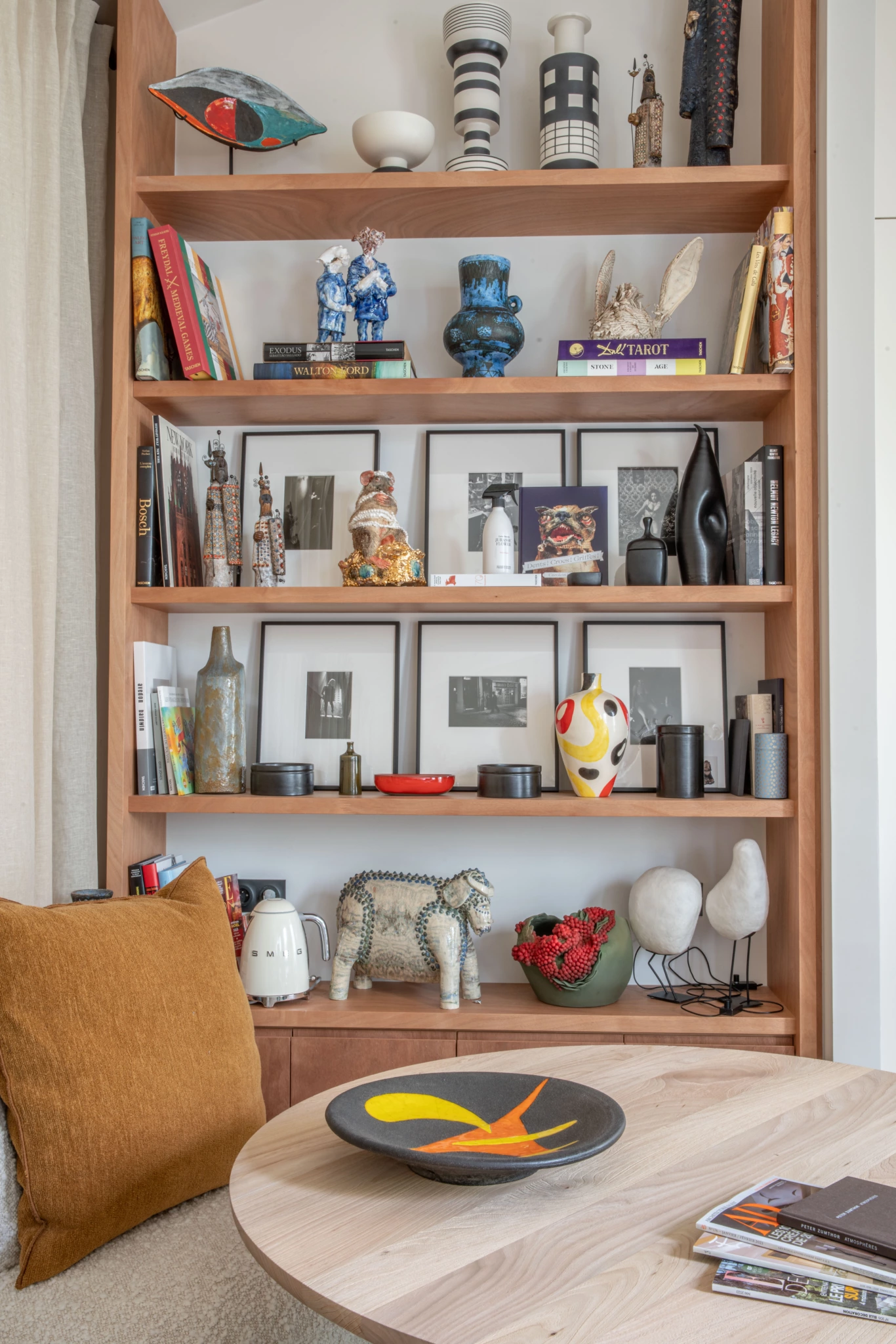 Espace de travail Paris 6ème 10 Etagère en bois avec livres, photos encadrées, poterie ; au premier plan, table ronde avec bol à motif d'oiseau ; coussin ocre sur la chaise de gauche.