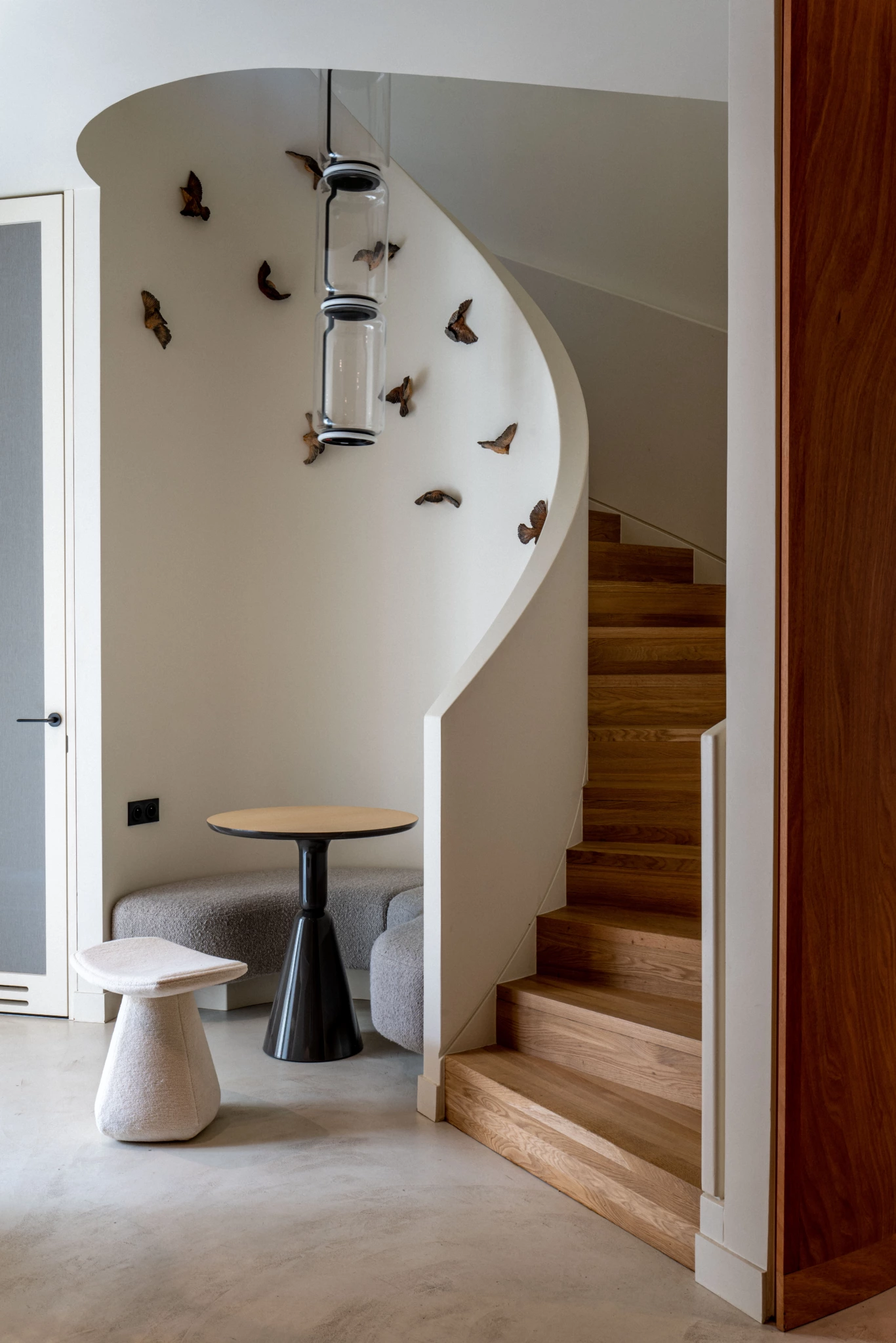 Espace de travail Paris 6ème Intérieur d'un studio photo avec escalier en bois incurvé, table ronde noire, tabouret blanc, art mural de l'oiseau volant, palette neutre et accents en bois.