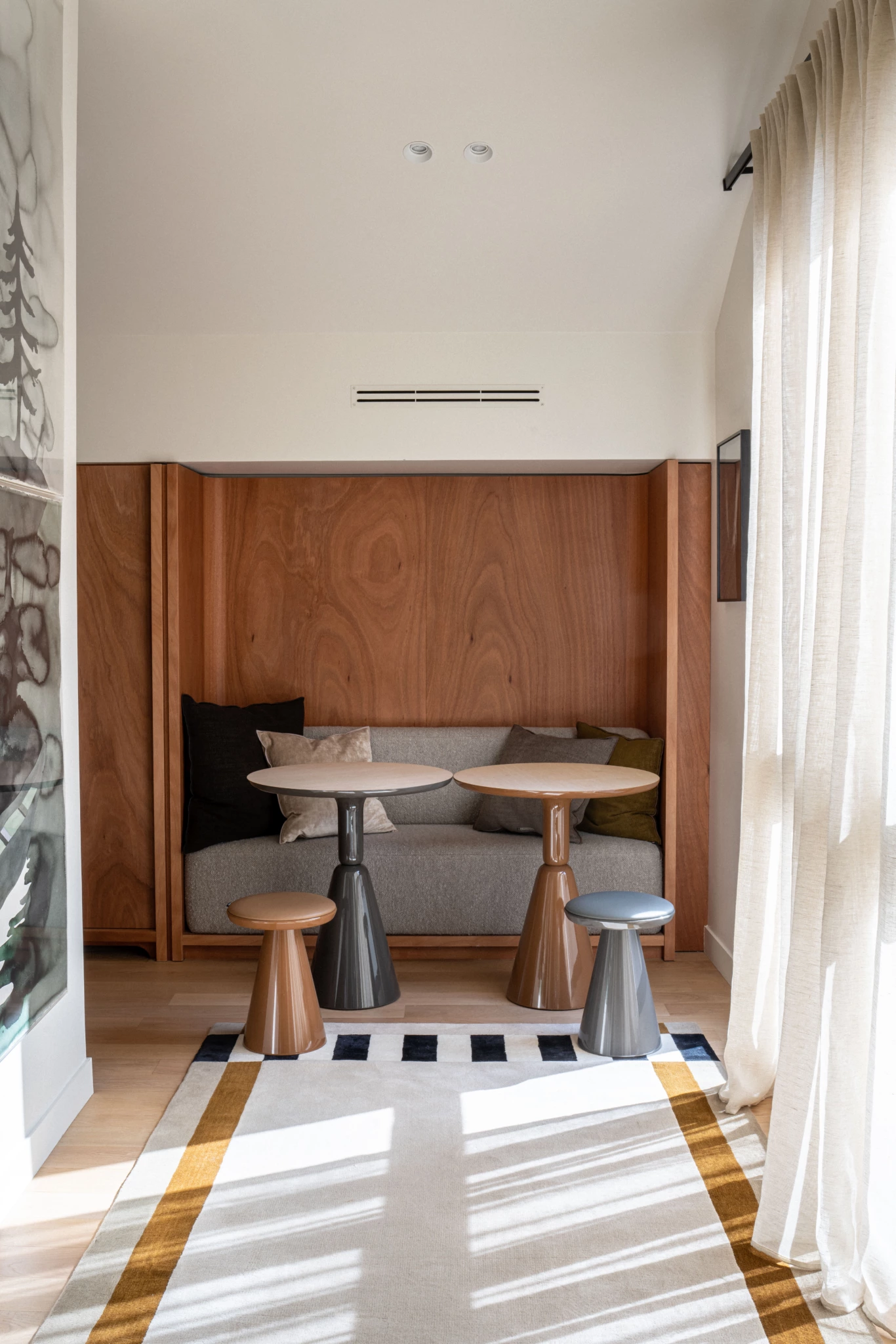 Espace de travail Paris 6ème 2 Coin en bois avec banc gris, deux tables rondes et trois tabourets ; la lumière naturelle à travers des rideaux diaphanes améliore la composition de l'espace.