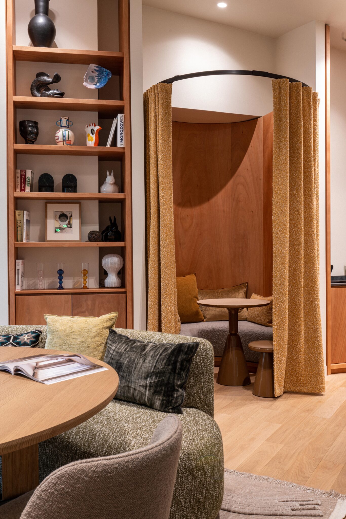 Espace de travail Paris 6ème 4 Coin salon avec table ronde et rideaux jaunes à côté d'étagères encastrées dans un espace de travail moderne, meublé de façon neutre, éclairage de studio.