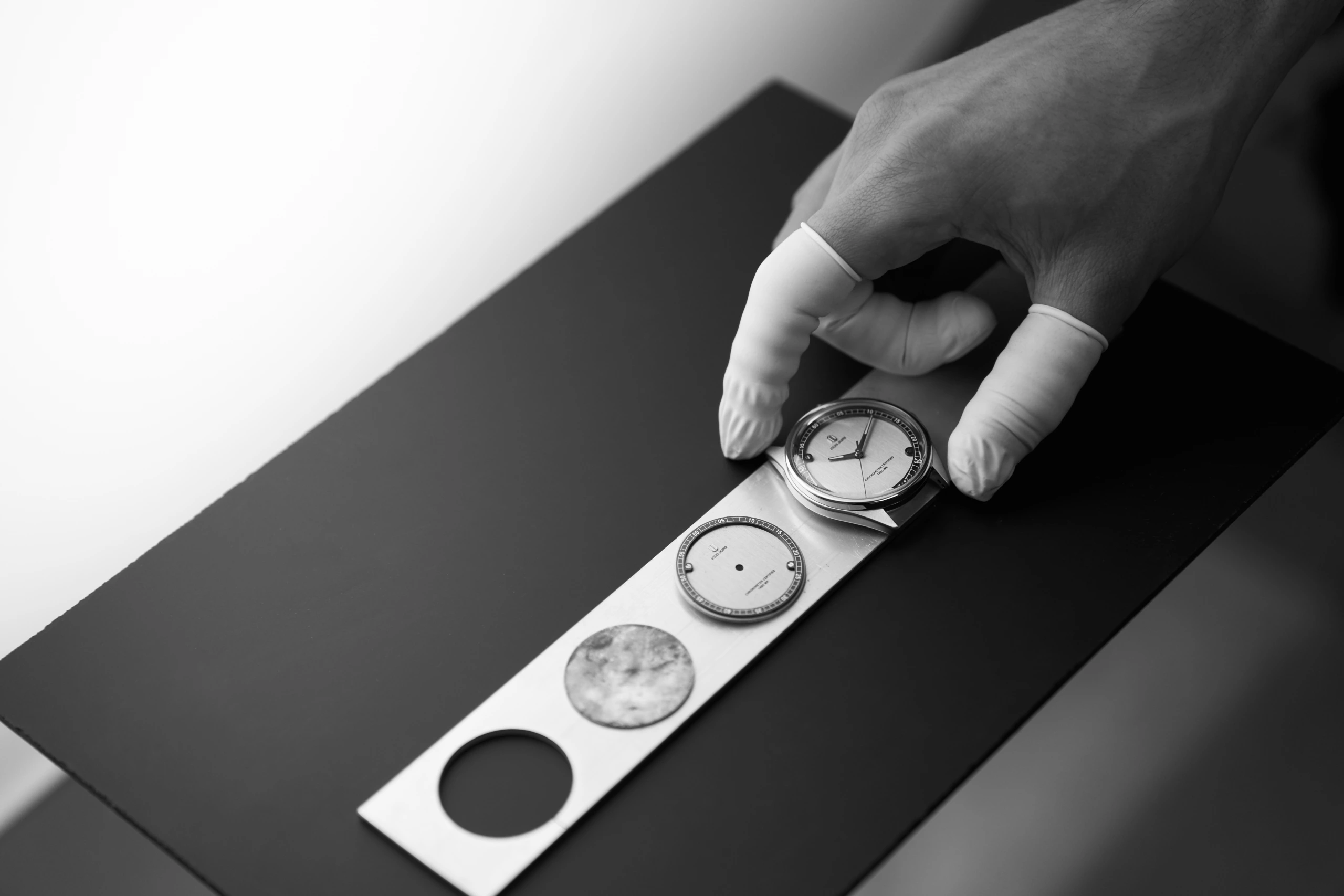 Une main gantée positionne un boîtier de montre-bracelet démonté au-dessus d'un plateau contenant des pièces sur une surface noire dans cette photo de studio en noir et blanc.