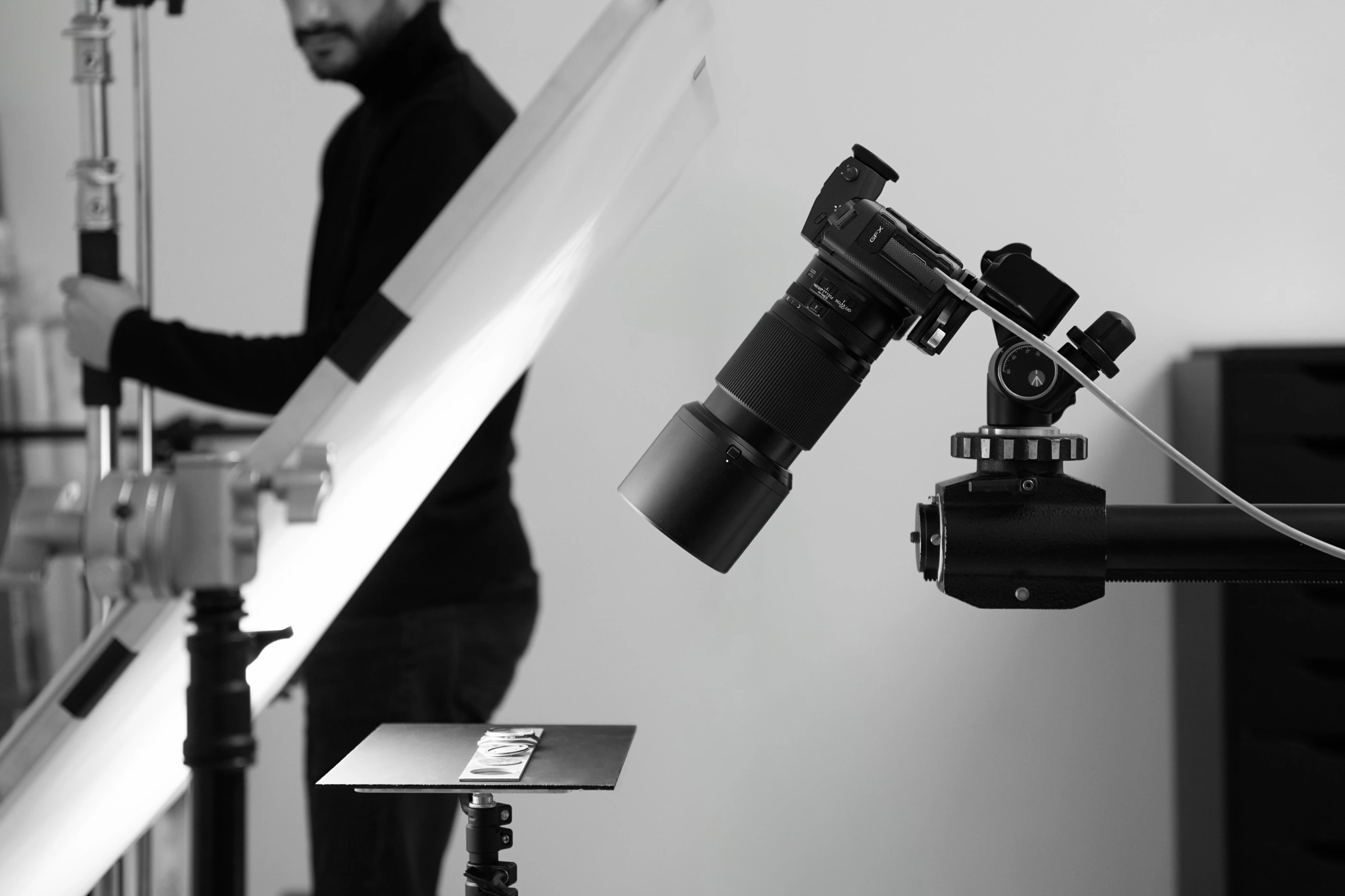 Gros plan d'une caméra orientée vers le bas sur un pied au-dessus d'une plate-forme de produits ; un technicien règle l'éclairage du studio en noir et blanc.
