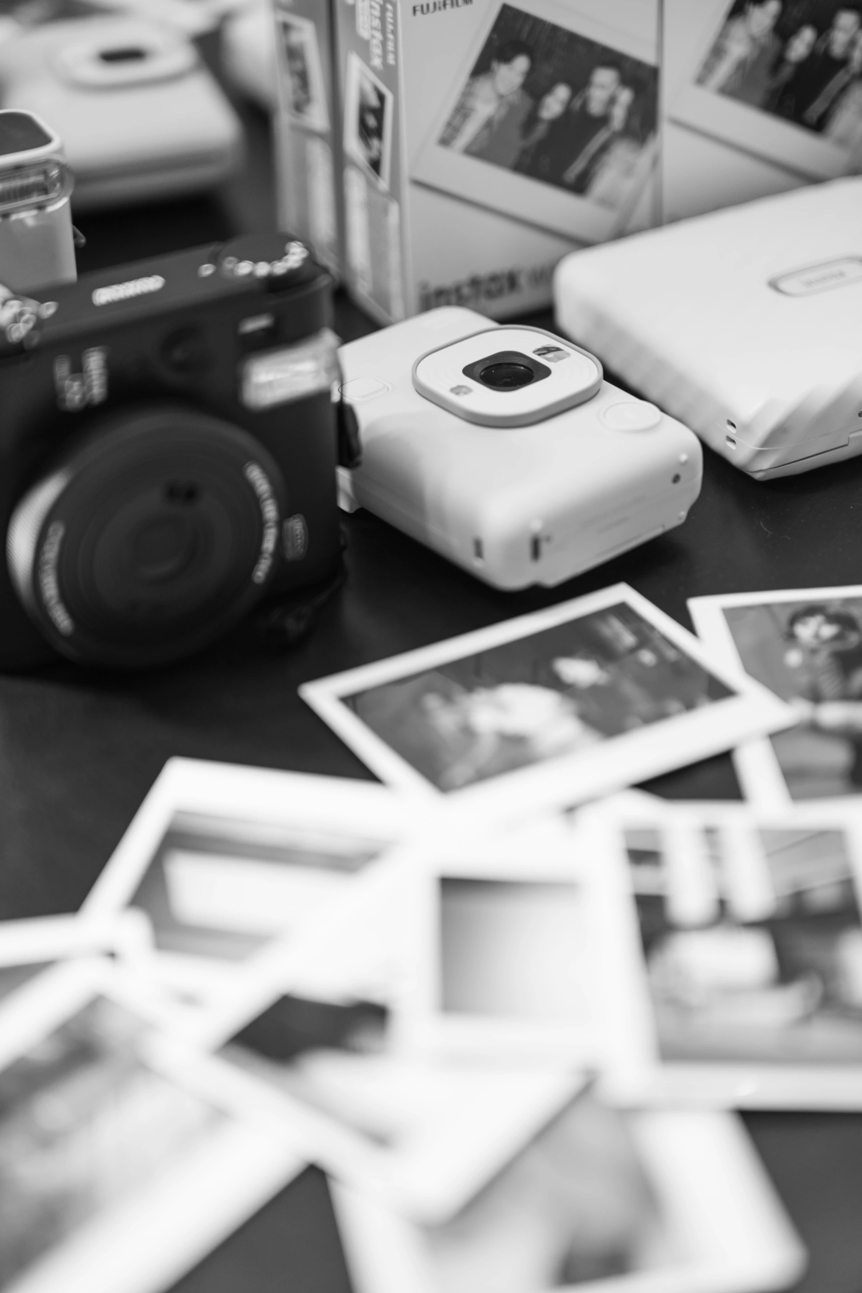 Image monochrome d'appareils photo instantanés, de boîtes de films et de tirages Argentique disposés sur une table avec des détails de photos flous.