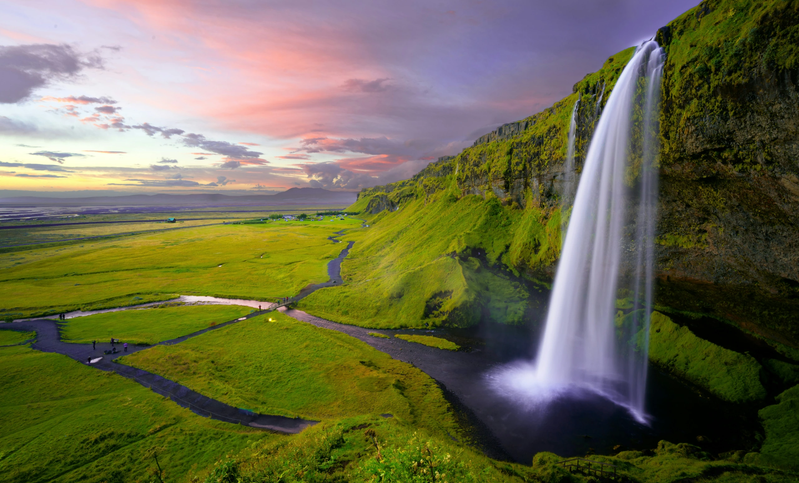 Une chute d'eau verticale s'écoule d'une falaise recouverte de mousse jusqu'à l'herbe en contrebas au coucher du soleil ; une rivière sinueuse et un ciel éclatant complètent la photo de paysage.
