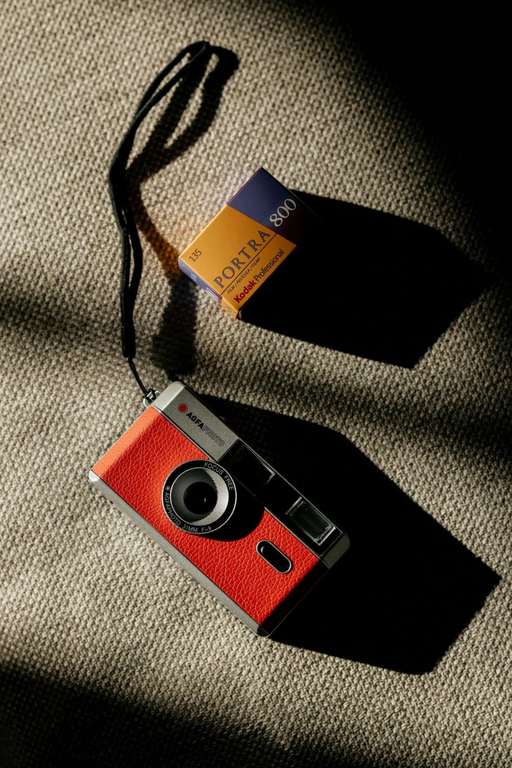 Un appareil photo compact avec un panneau frontal rouge et une courroie noire à côté d'un film Kodak Portra 800 sur une surface beige, en lumière naturelle.
