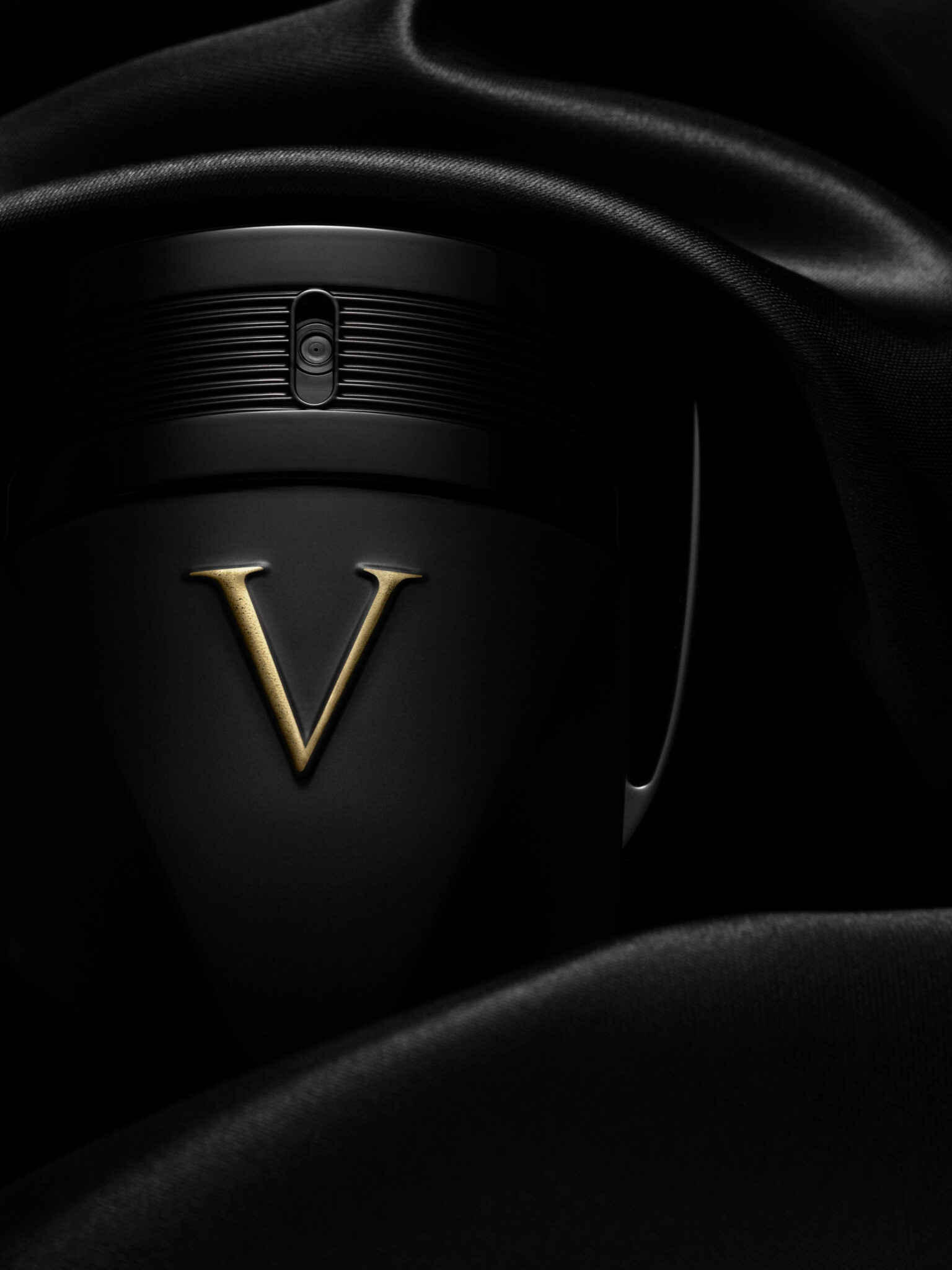 Invictus_Victory_Absolu Un récipient noir avec un V doré est partiellement recouvert de satin noir, photographié sous un éclairage dramatique pour mettre en valeur la texture et les détails.