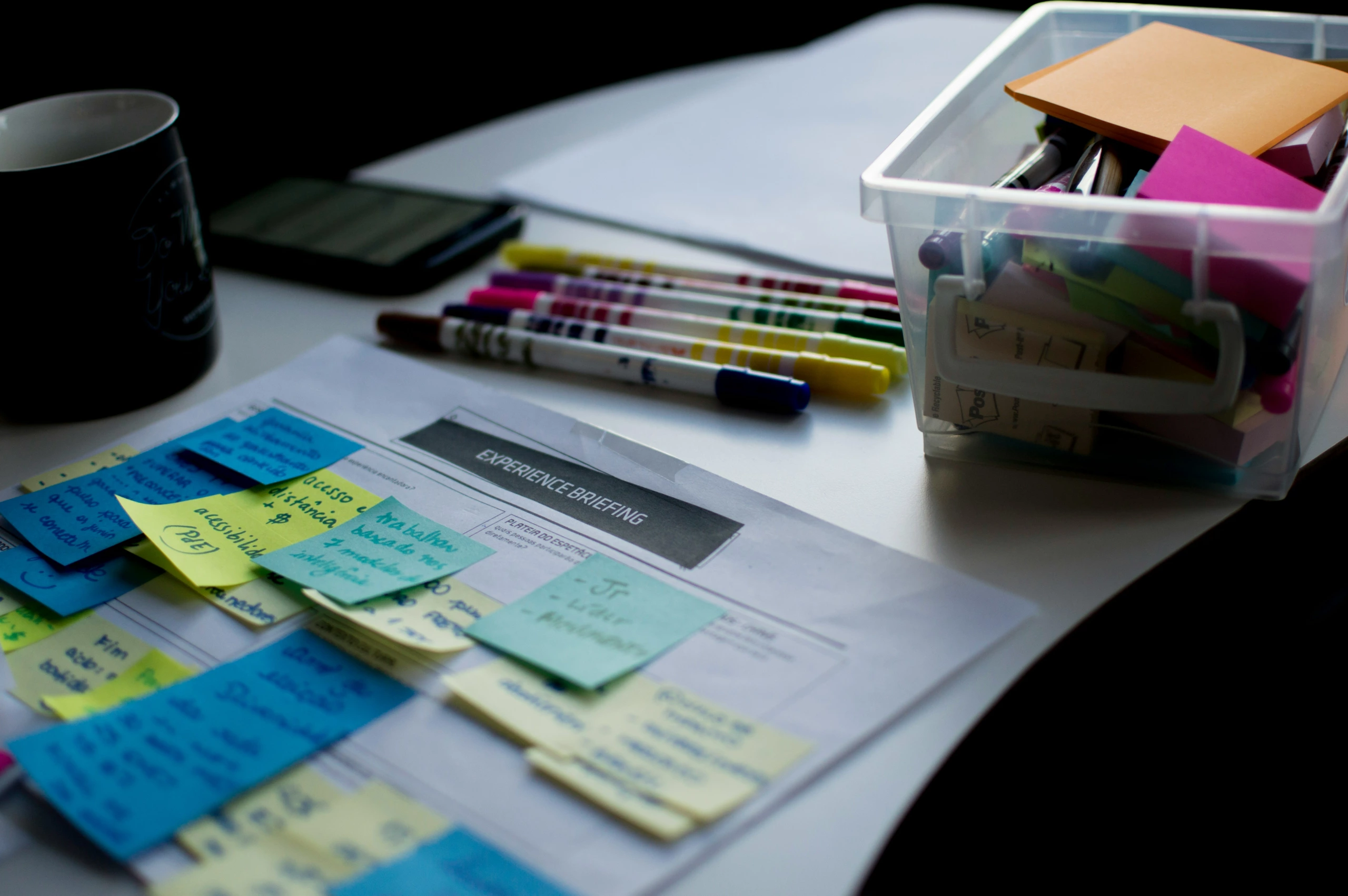 Espace de travail bien éclairé d'un studio de photographie avec un bureau organisé, des documents étiquetés avec des notes autocollantes, des marqueurs, des stylos, un téléphone et une tasse à café visible.
