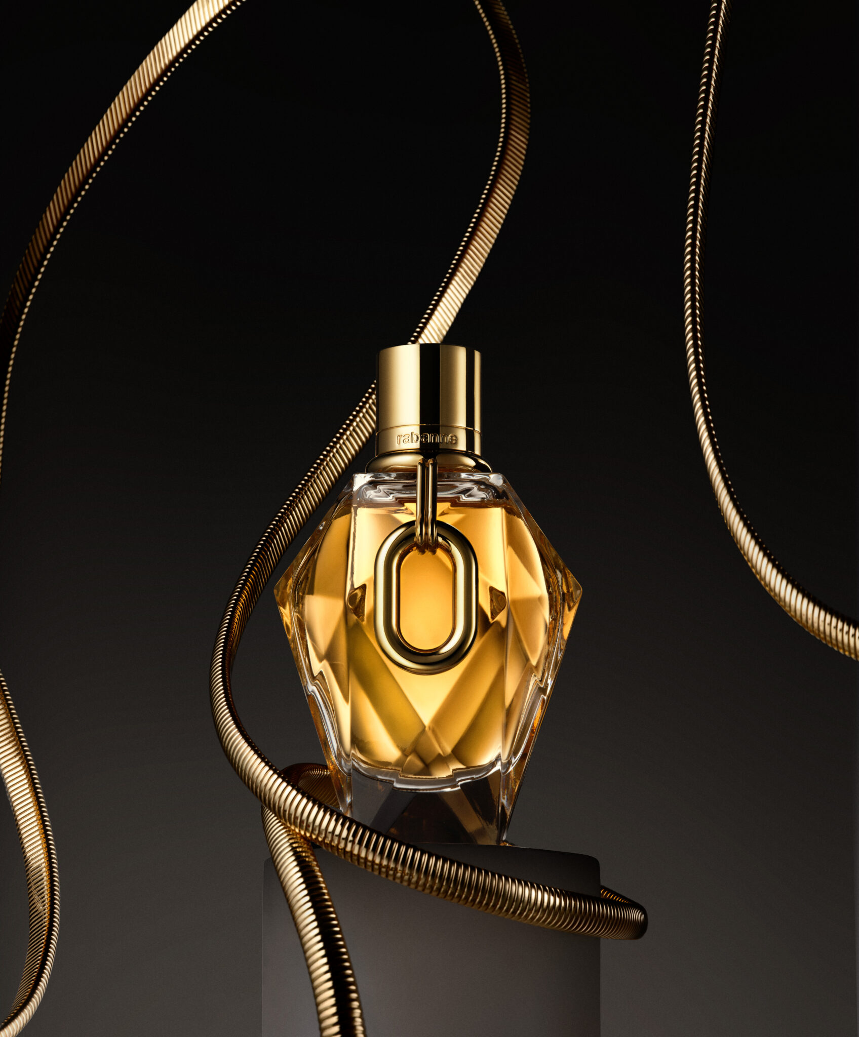 Million Gold For Her Un flacon de parfum en verre facetté avec des détails en or et une bague en or est posé sur un piédestal, encadré par des câbles métalliques sur un fond de studio sombre.