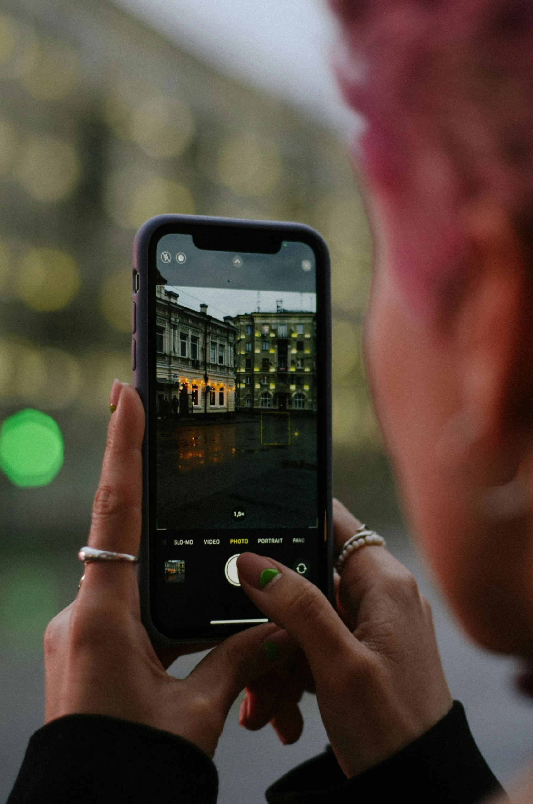 Un photographe aux cheveux roses capture des bâtiments historiques illuminés au crépuscule à l'aide d'un smartphone, l'interface de l'appareil photo étant visible à l'écran.