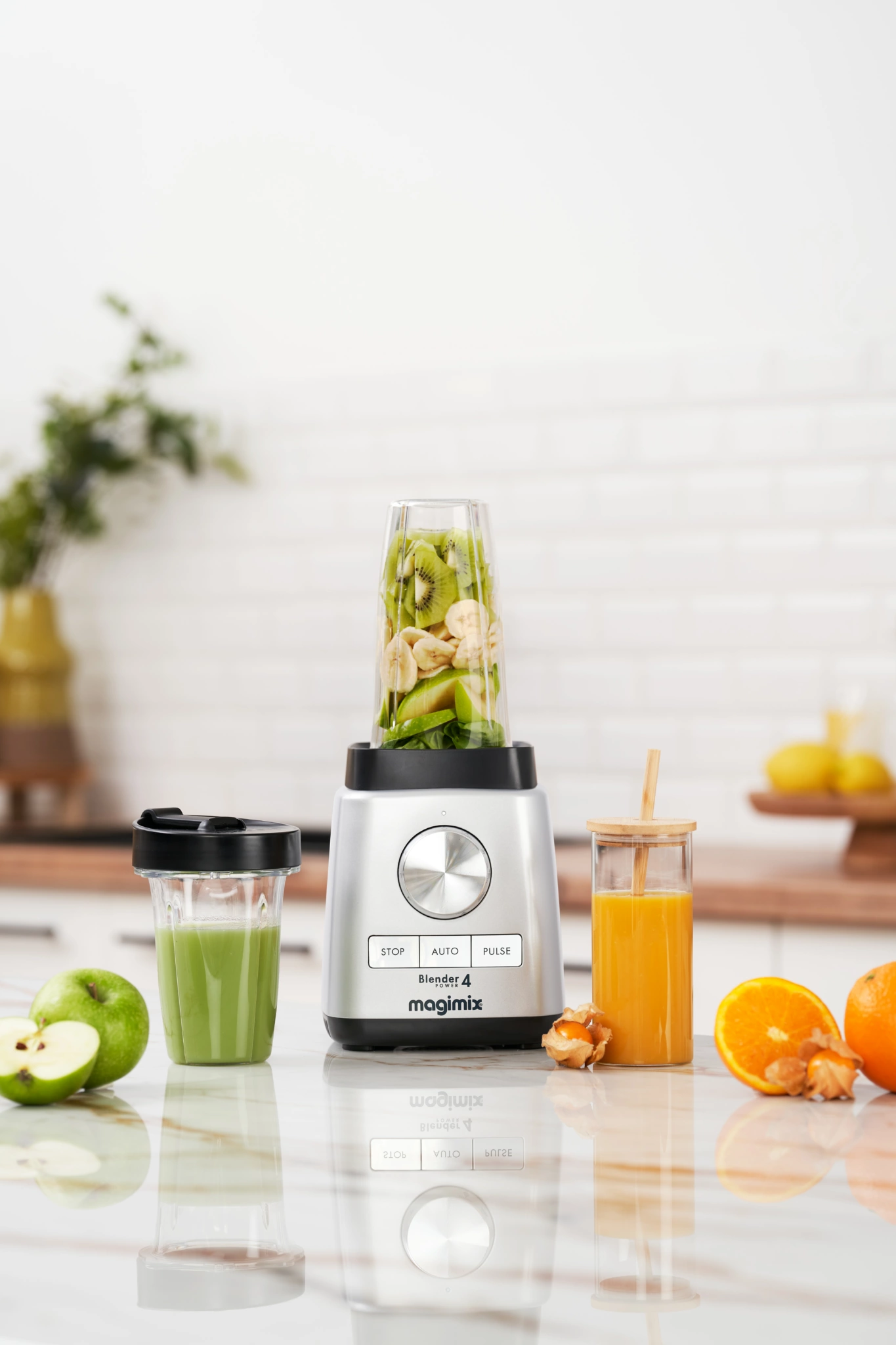 MAGIMIX – 20 Photo publicitaire d'un mixeur Magimix rempli de tranches de kiwi et de banane, posé sur un plan de travail de cuisine à côté d'un gobelet à smoothie et d'un verre de jus d'orange.