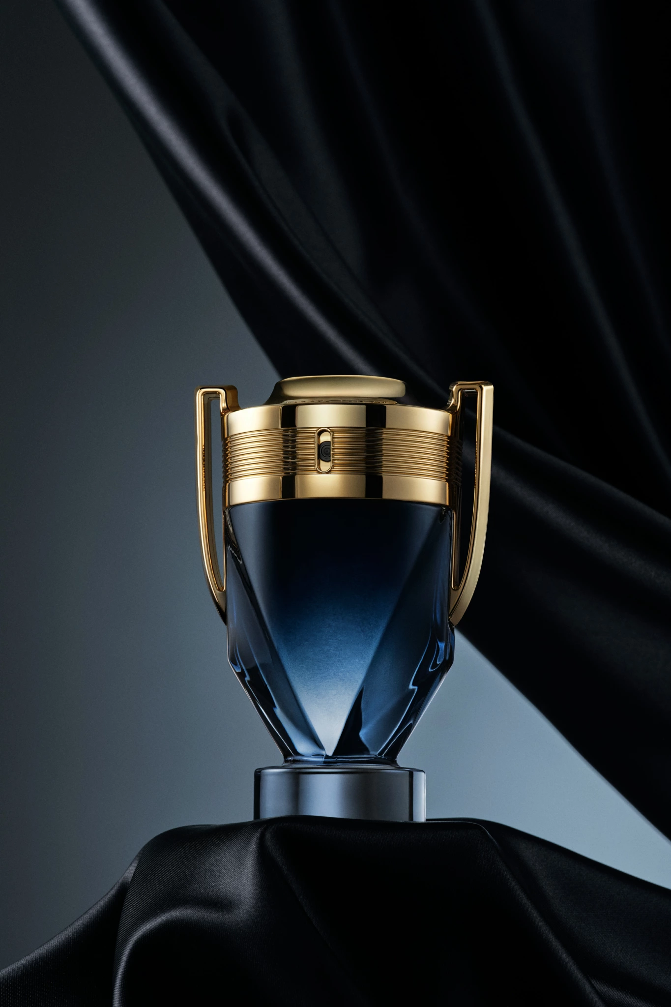 Invictus_Parfum_2 Photographie Flacon de parfum avec bouchon doré, verre bleu à facettes et poignées sur une surface noire ; toile de fond en tissu noir pour la photographie de produits.
