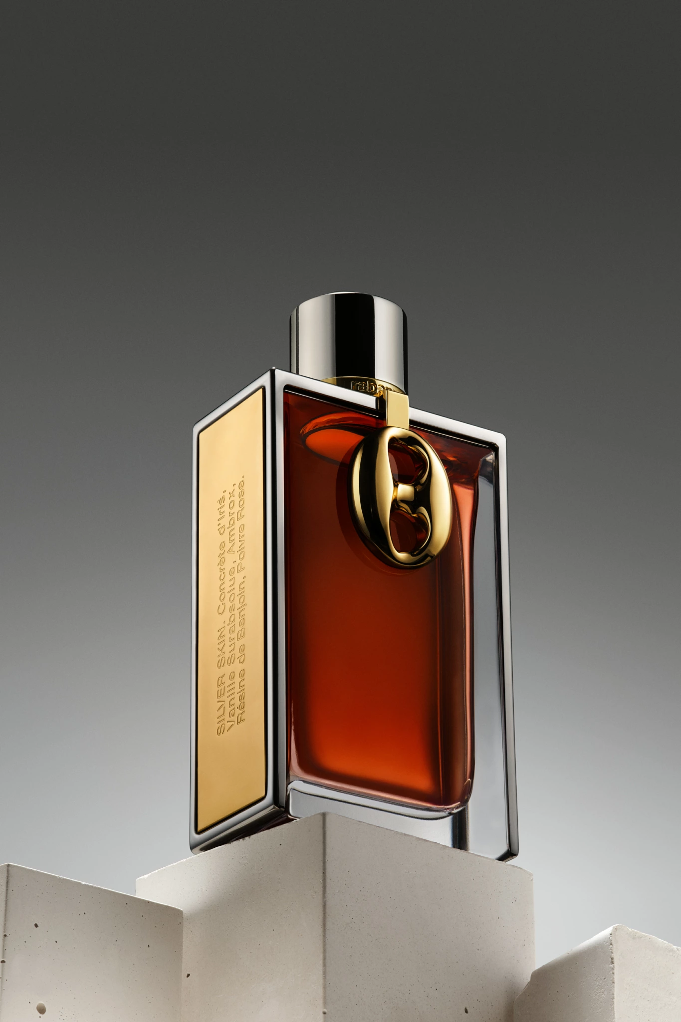 SILVER_SKIN Flacon de parfum rectangulaire avec un liquide ambré, une plaque latérale dorée et un accent "B", mis en scène sur des blocs géométriques blancs dans un studio.