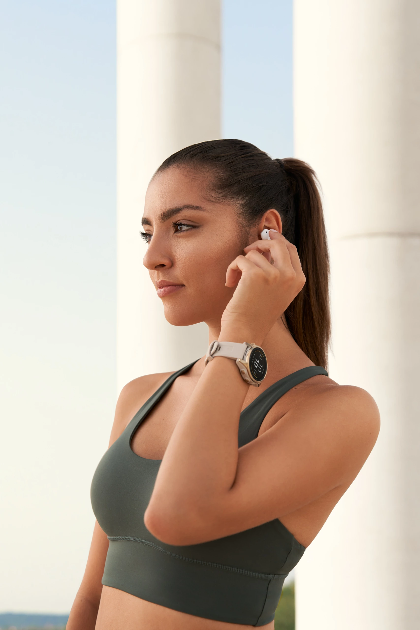 Une femme avec une queue de cheval, vêtue d'un soutien-gorge de sport vert foncé et d'une smartwatch, ajuste un écouteur en plein air, avec des colonnes blanches et le ciel dans le cadre.