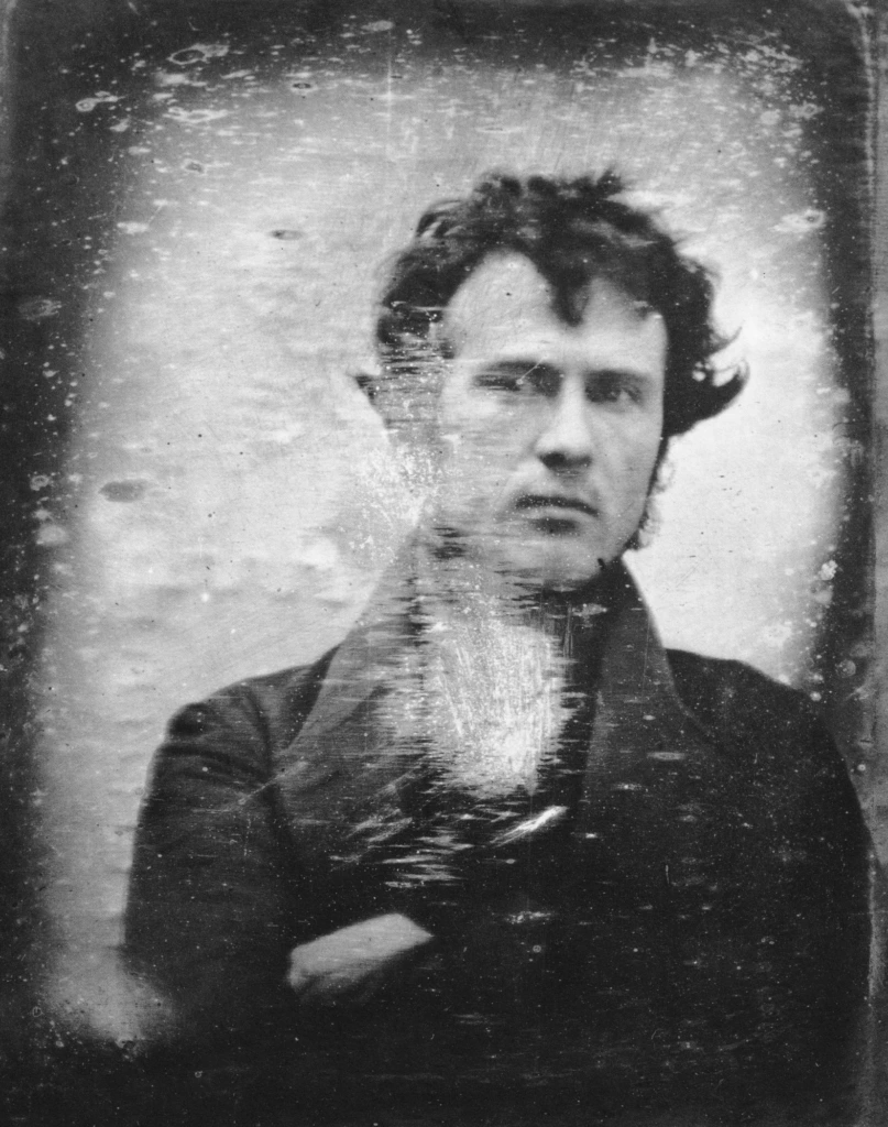 Portrait d'archive en noir et blanc représentant un homme aux cheveux bouclés, à l'expression neutre, vêtu d'un manteau foncé et les bras croisés. La photographie présente des traces d'usure dues à son âge, notamment des rayures superficielles et des signes visibles de détérioration.