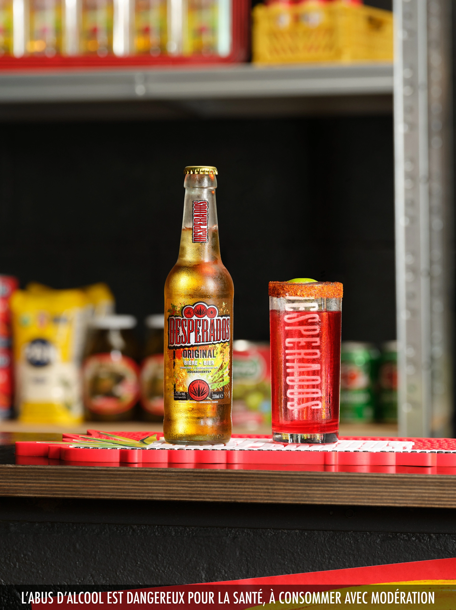Desperados_LaDespenida_01 Cette composition réalisée en studio met en scène une bouteille de bière Desperados et un cocktail rouge au bord salé, disposés sur le comptoir d'un bar. À l'arrière-plan, on aperçoit des étagères remplies de snacks et de bouteilles, rendues légèrement floues grâce à des techniques de profondeur de champ.
