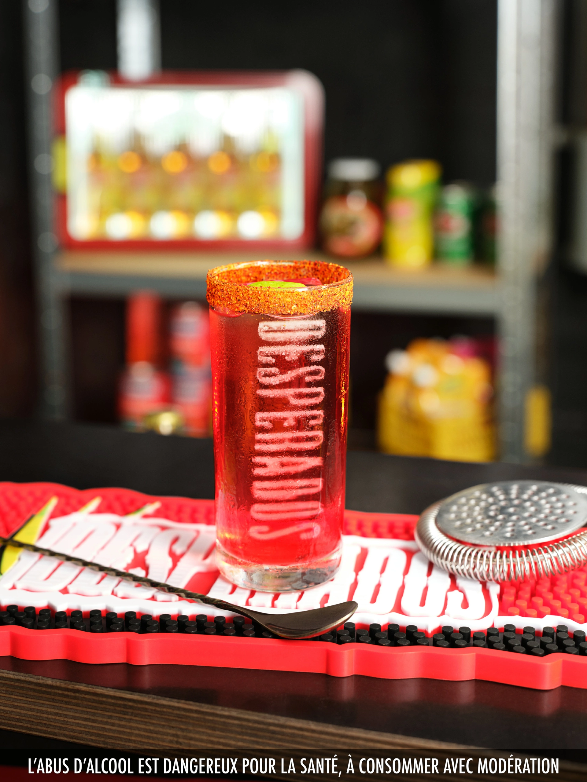 Desperados_LaDespenida_02 Un grand verre transparent contenant un liquide rouge, dont le bord est enduit d'orange et portant l'inscription « Desperados », est posé sur un tapis de bar à l'effigie de la marque. À côté du verre se trouvent une passoire à cocktail en métal et une cuillère à mélange, tandis que d'autres accessoires de bar apparaissent légèrement flous à l'arrière-plan. Un texte d'avertissement français sur les risques liés à l'alcool est affiché le long du bord inférieur de l'image. La composition illustre une maîtrise efficace de la profondeur de champ en photographie de produit.