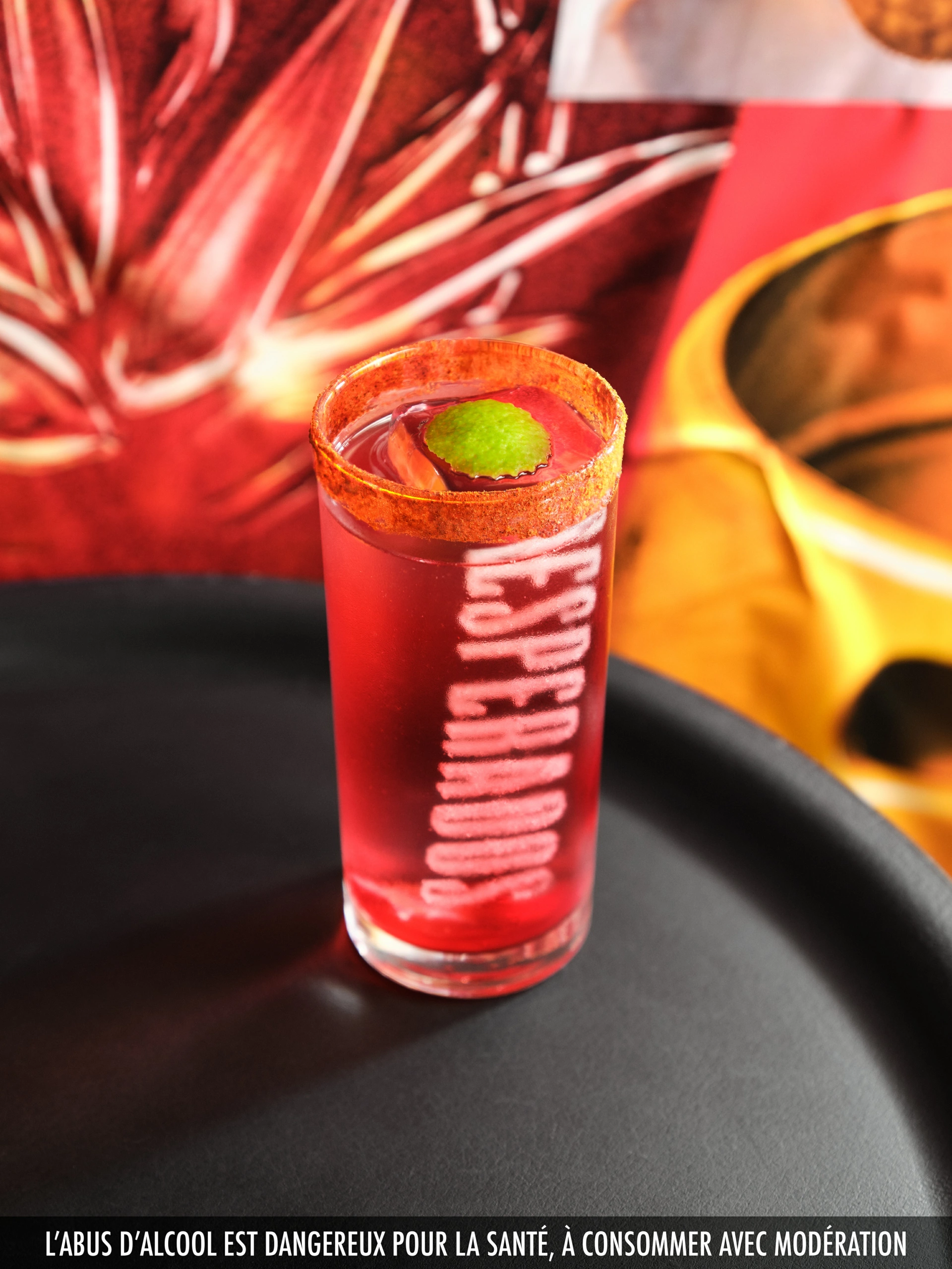 Desperados_LaDespenida_04 Un verre transparent contenant un cocktail rouge, dont le bord est saupoudré de poudre de piment et orné d'une feuille verte, est posé sur un plateau de table noir. Le mot « ESPERADOS » est visible sur le verre. L'arrière-plan présente des motifs abstraits rouges et jaunes, créant un effet d'éclairage de studio maîtrisé pour la photographie de produit.