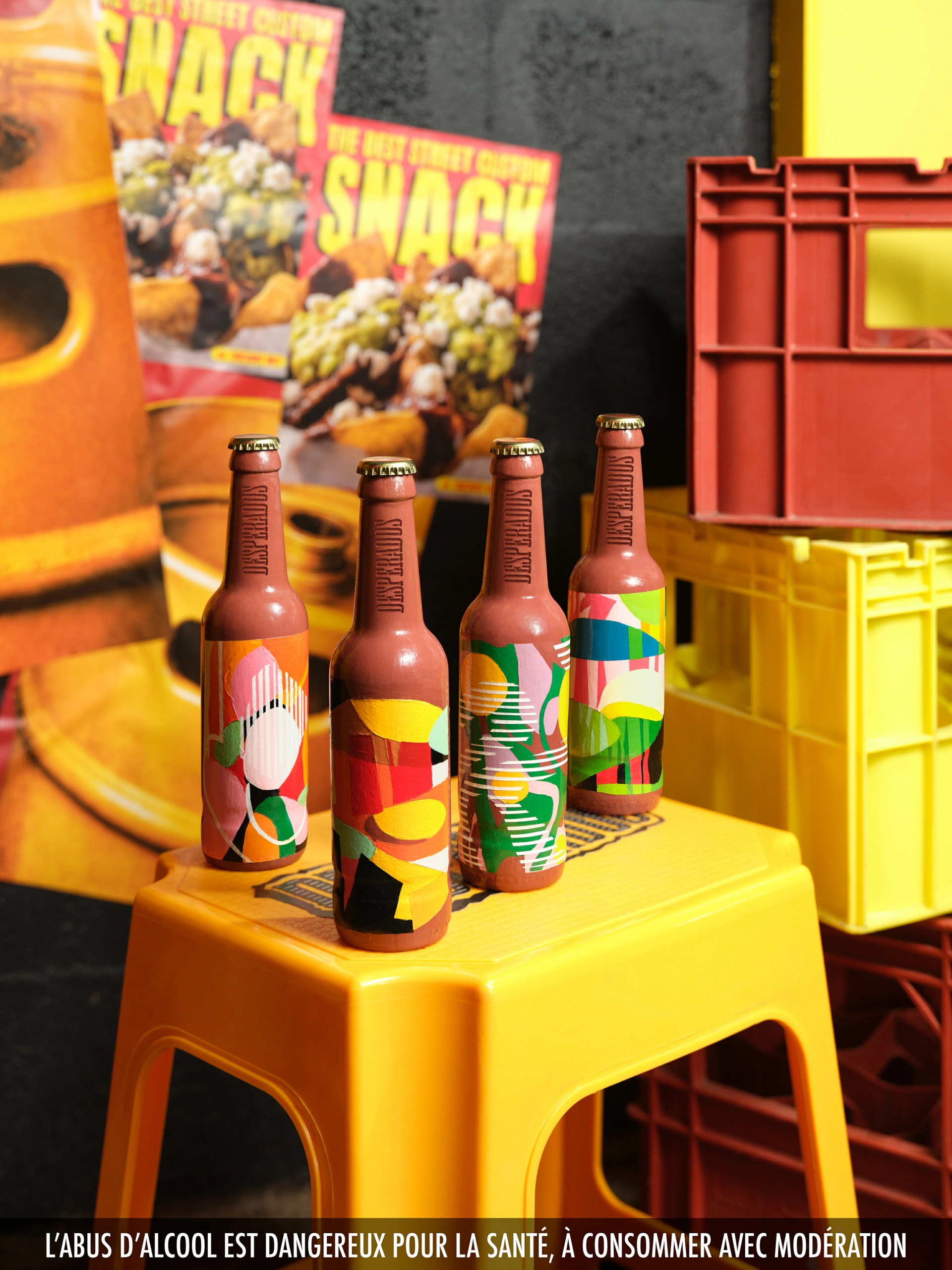 Desperados_LaDespenida_05 Une photo de produit en haute résolution montre quatre bouteilles de bière aux étiquettes distinctes et abstraites, disposées sur un tabouret en plastique jaune. La composition comprend des caisses en plastique empilées et deux magazines intitulés « SNACK » à l'arrière-plan. Un avertissement français sur les risques liés à la consommation d'alcool apparaît au bas de l'image.