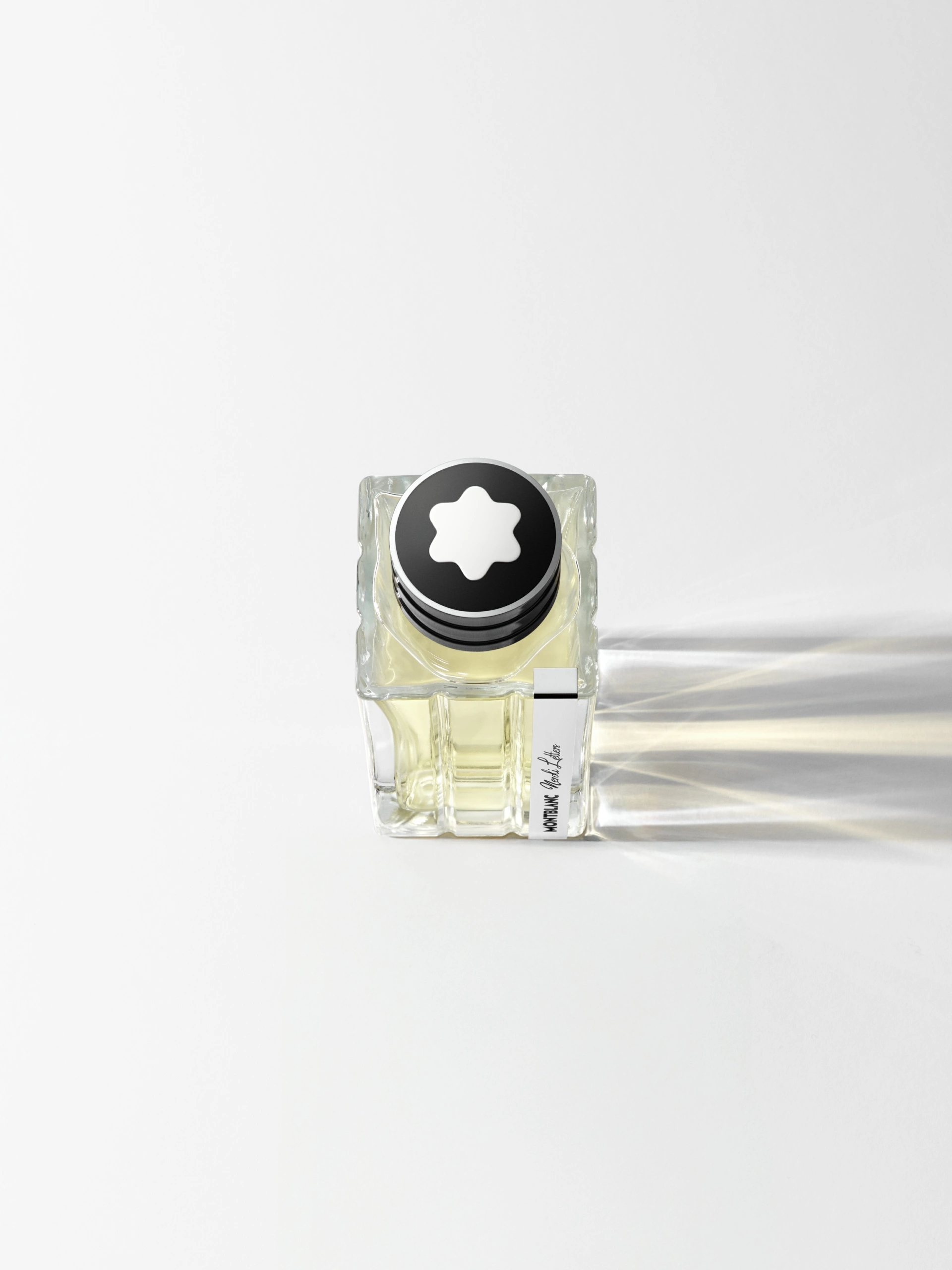 SOLO_MONTBLANC_FOCUS_BOTTLE_R3 Vue de dessus d'une bouteille carrée en verre transparent contenant un liquide jaune. La bouteille est munie d'un bouchon noir de forme circulaire orné d'un emblème blanc en forme d'étoile. Un éclairage latéral doux crée des ombres bien définies à droite sur un fond blanc épuré, illustrant ainsi une technique efficace de photographie de produit.